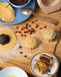 Mooncakes with floral designs, red bean paste, and dough are arranged on a wooden surface with a spoon, mooncake mold, and scattered red beans. A sliced mooncake is on a decorative plate.