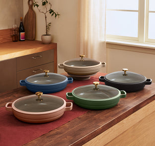 Five ceramic casserole dishes in different colors with glass lids and gold handles are displayed on a kitchen counter with a red table runner.