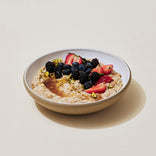A bowl of oatmeal topped with strawberries, blackberries, blueberries, chopped nuts, and a drizzle of syrup, on a light background.
