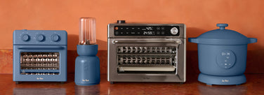 Collection of kitchen appliances including a toaster oven, blender, air fryer, and slow cooker on a wooden surface with an orange wall background.