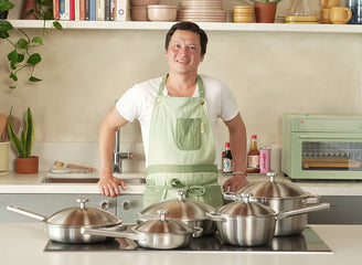 Man in a kitchen wearing a green apron with stainless steel pots and pans on the stove.