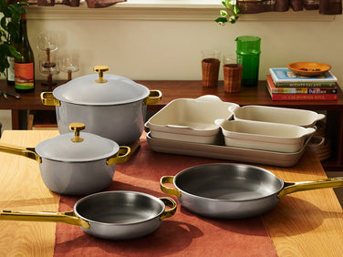 A coordinated cookware set arranged on a wooden table in a kitchen setting, including two gray pots with gold-tone handles and knobs, two gray pans with gold-tone handles, a stack of cream ceramic baking dishes, and a gray roasting pan. Books, glasses, and a plant are visible in the background.