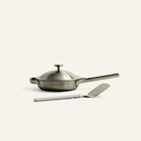 Stainless steel frying pan with lid and spatula on a light beige background
