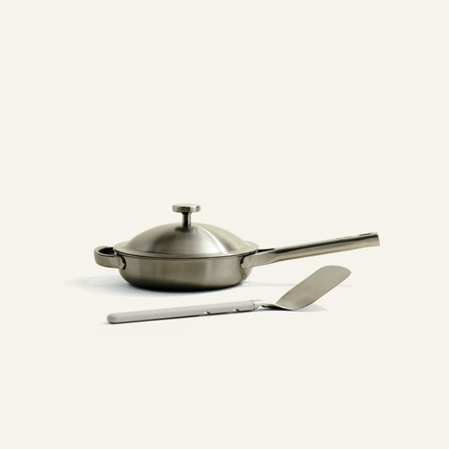 Stainless steel frying pan with lid and spatula on a light beige background