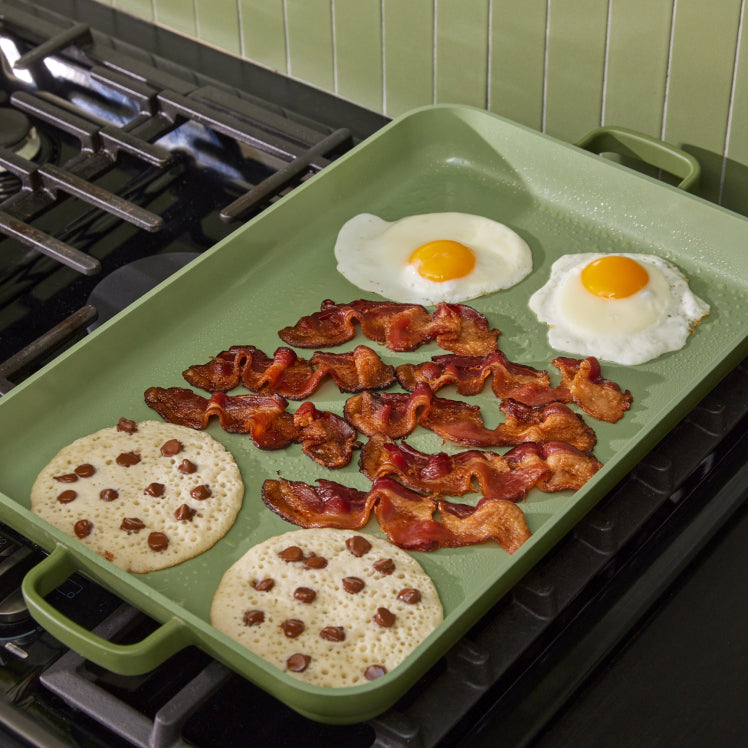 green griddle pan on stove cooking pancakes, bacon, and eggs