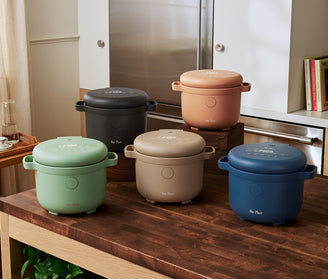 Five colorful rice cookers are displayed on a wooden kitchen island, with a modern kitchen setup in the background.