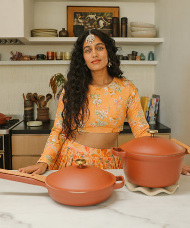 A woman in a yellow floral outfit stands in a kitchen behind a skillet and Dutch oven on a marble countertop.