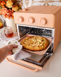 A hand with an oven mitt pulls a tray of freshly baked apple dessert from a peach-colored countertop oven. Flowers and a jar of honey are visible in the background.