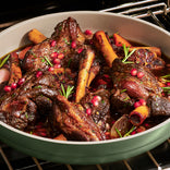 Braised lamb shanks in a pan with rosemary, pomegranate seeds, and sliced garlic, cooked in a dark, rich sauce.