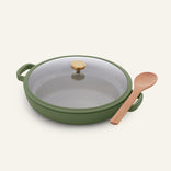 Versatile Dual Handle Always Pan in green, featuring a nonstick surface, convenient side handles, a glass lid with a gold knob, and accompanied by a wooden spoon on a light background—a non-toxic addition to any modern kitchen.