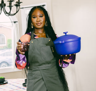 Woman in an apron holding a blue pot and a glass of red beverage stands in a dining room near a table and window.