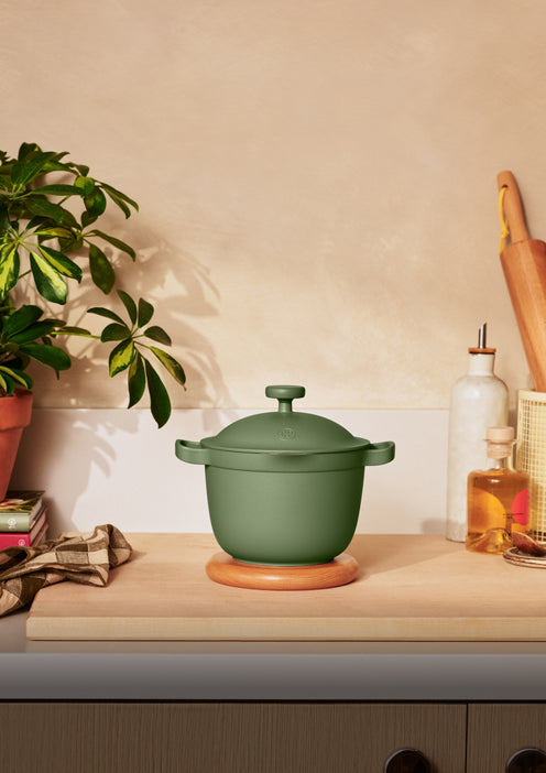 A green covered pot sits on a wooden trivet on a kitchen counter, surrounded by a plant, kitchen towel, rolling pin, and bottles of oil.