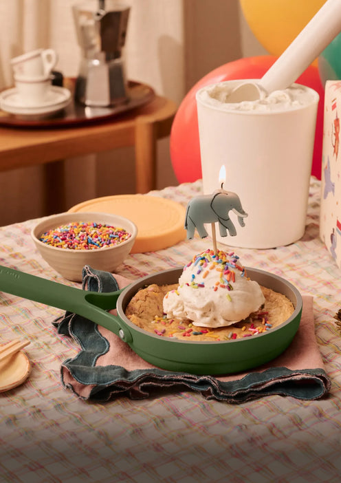 A skillet cookie topped with ice cream, sprinkles, and an elephant-shaped candle sits on a table next to a bowl of sprinkles, a container of marshmallows, and balloons in the background.