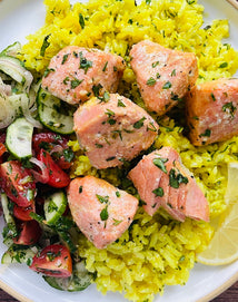 Plate with yellow rice, herb-seasoned salmon chunks, sliced cucumber and tomato salad, and a lemon wedge.