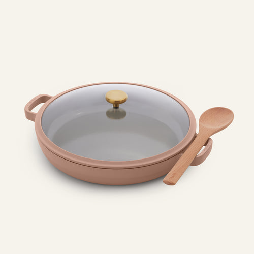 The Dual Handle Always Pan is a round, light brown nonstick pan featuring two handles, a clear glass lid with a gold knob, and a wooden spoon resting on the right handle, all set against a plain background.