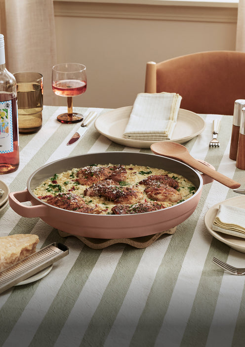 A table set with a skillet of baked chicken and vegetables, a glass of rosé wine, plates, cutlery, and cloth napkins on a striped tablecloth.