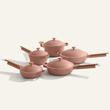 The Cookware Set+ is a 16-piece ceramic collection with four pink nonstick pots and pans, matching lids, and wooden handles, displayed on a light background.