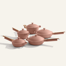 The Cookware Set+ is a 16-piece ceramic collection with four pink nonstick pots and pans, matching lids, and wooden handles, displayed on a light background.