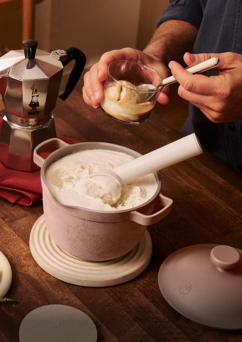 A person scoops vanilla ice cream from a pot into a glass cup next to a moka pot on a wooden table.