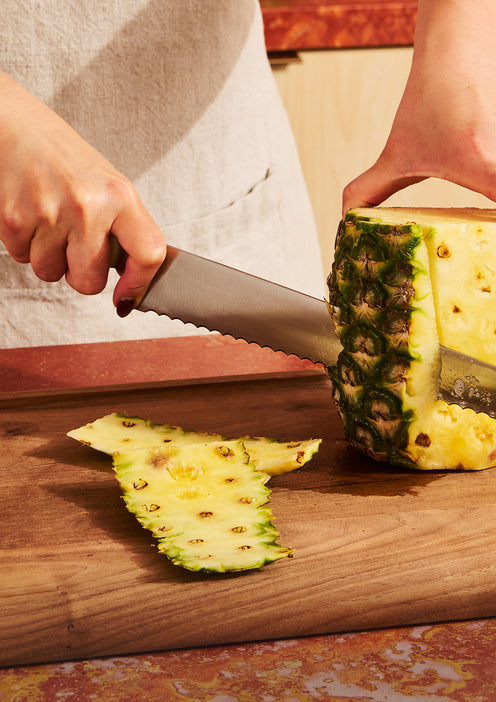 A person uses a serrated knife to cut the skin off a pineapple on a wooden cutting board.