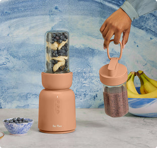 A hand holds a smoothie jar with a handle next to a blender filled with blueberries and banana slices; bananas and a bowl of blueberries sit nearby.