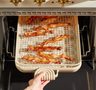 A hand pulls out an oven rack holding a tray with cooked bacon strips on a wire mesh baking sheet.