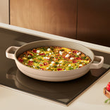 A round ceramic pan with handles holds a baked frittata topped with herbs and cheese, sitting on a stovetop in a modern kitchen.
