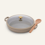 The Dual Handle Always Pan in beige features a ceramic nonstick surface, two handles for easy maneuvering, a glass lid with a gold knob, and comes with a wooden spoon resting beside it on a light background; this versatile, non-toxic cookware is perfect for everyday use.