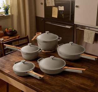 Five pieces of light gray cookware with wooden handles are arranged on a wooden kitchen table in a modern kitchen setting.