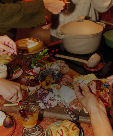 A table set with various dishes including a cake, spreads, fruit, and two large covered pots, with people standing nearby.