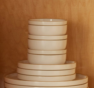 A stack of cream-colored ceramic bowls and plates with a thin gold rim, arranged neatly against a warm wood-grain background.