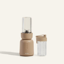 A beige countertop blender with a clear blending jar attached and a matching clear travel cup with a lid, both on a neutral background.
