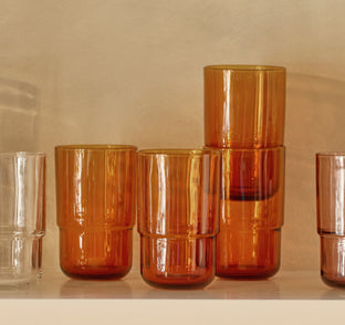 Six amber-colored glass tumblers and one clear tumbler are arranged on a shelf against a beige background.