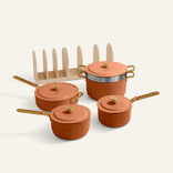 The Essentials Saucepan Set includes four orange pots and pans with lids, gold handles, a beige rack organizer, and a non-toxic nonstick coating, all displayed on a light background.