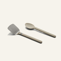 A stainless steel spatula and a tan serving spoon from a Titanium Pro Utensil Set lying side by side.