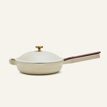 The Always Pan 2.0 is a beige, nonstick frying pan from a bestselling cookware system, featuring a matching domed lid, gold knob, and long handle on a plain light background.