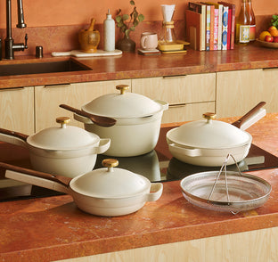 A set of four cream-colored pots and pans with lids and wooden handles on a kitchen counter, alongside a metal strainer basket.