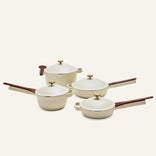 The Cookware Set features four cream-colored pieces inspired by the Always Pan and Perfect Pot, with wooden handles and brass knobs: two frying pans, a saucepan with lid, and a pot with lid, displayed on a neutral background.