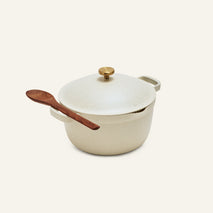 The Perfect Pot is a cream-colored, family-sized ceramic pot with a lid and gold knob, featuring a wooden spoon resting on its side handle, set against a plain background.