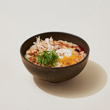 A dark bowl filled with rice porridge topped with shredded chicken, a soft-boiled egg, sliced green onions, and seasonings.
