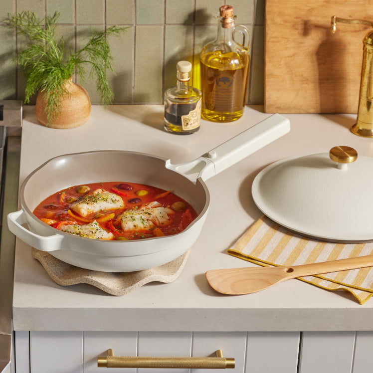 A white Always Pan with a tomato-based fish dish on a counter with a lid, spatula, and olive oil nearby